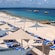 An area of beach covered in lounge chairs and shading tarps near a pier, boats, a building and palm trees